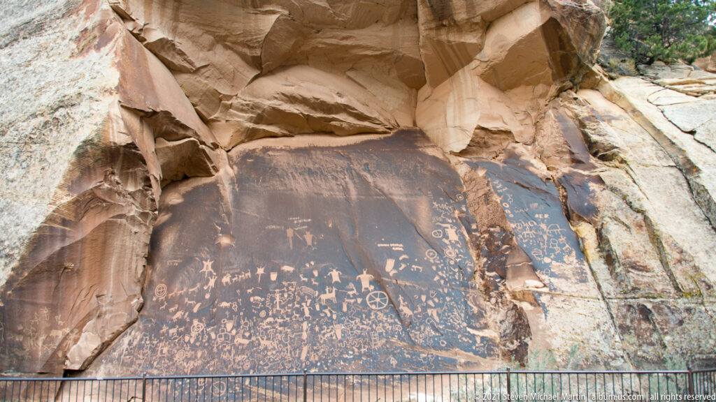 A dark petroglyph panel fills the base of a sandstone alcove, framed by pale ledges and a protective railing.