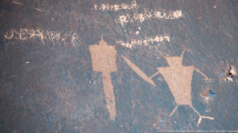 Two large anthropomorphic petroglyphs with extended limbs appear on dark sandstone, with a diagonal line between them.