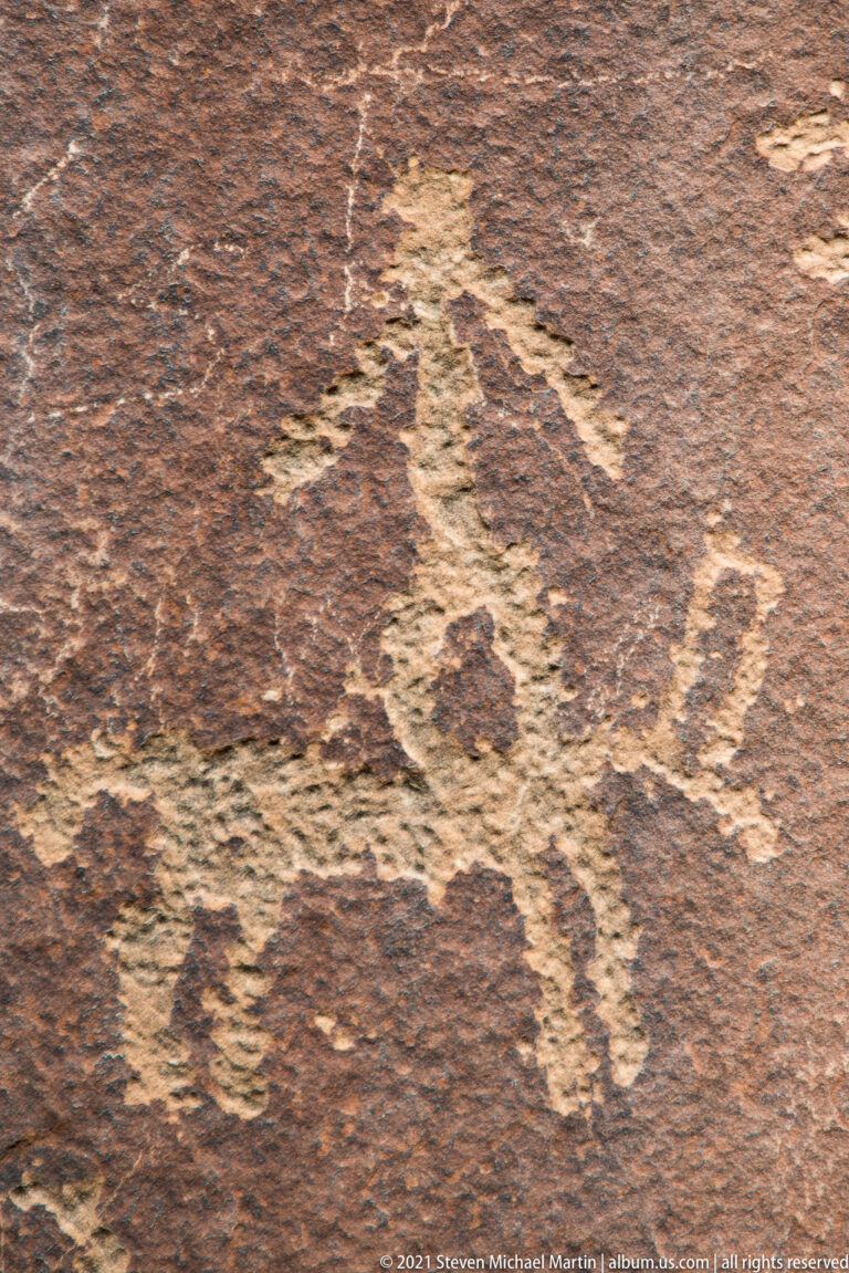 A broad pale animal-like figure with short legs and curved extensions is pecked into dark sandstone.
