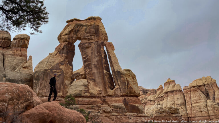 A hiker stands on a red boulder beneath the tall supports and lintel of Druid Arch.