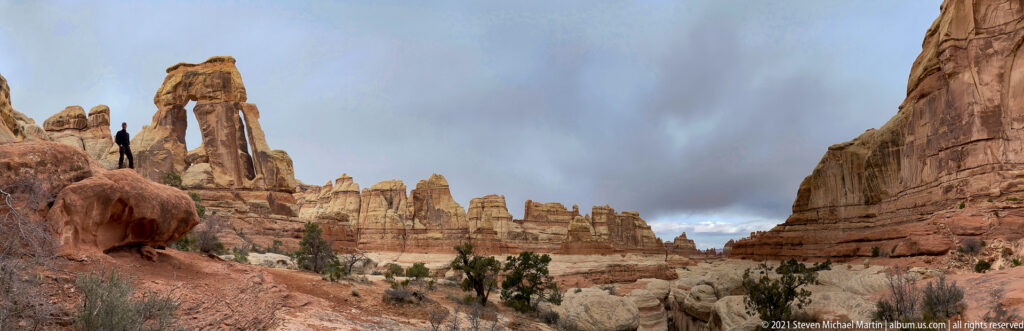 Druid Arch stands at left across an open rocky approach, balanced by a narrow pinnacle at right.