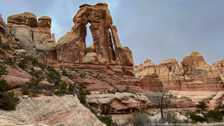 A rounded foreground boulder leads toward Druid Arch and pale sandstone walls under blue sky.