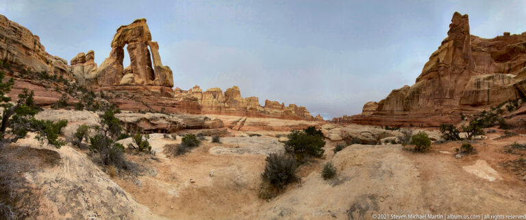 A sandy trail approaches Druid Arch from a shrub-lined foreground beneath layered sandstone walls.