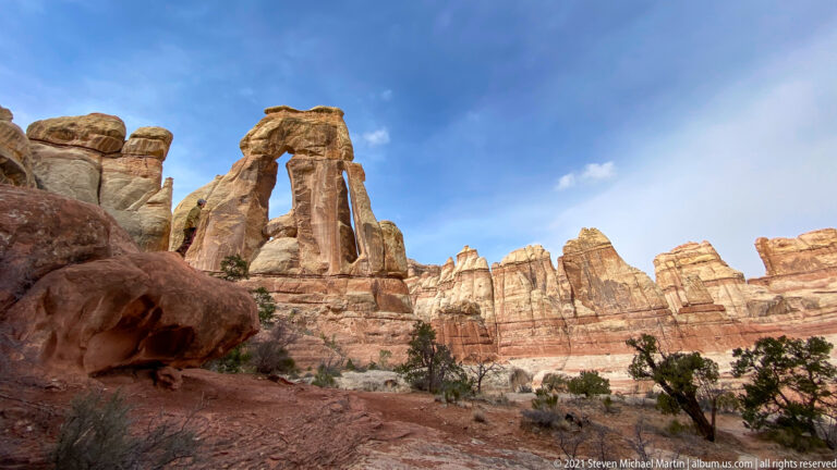 A red boulder fills the foreground below Druid Arch, where a small hiker stands among pale rock walls.