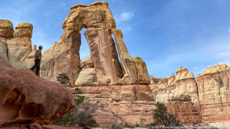A hiker stands at the base of Druid Arch beside its tall sunlit sandstone columns.