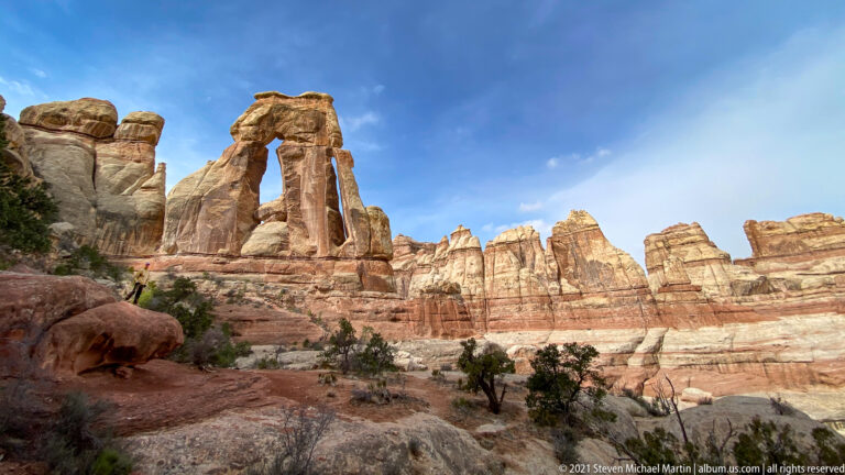 Druid Arch appears at far left across a broad basin, with a tiny hiker below and sandstone walls beyond.