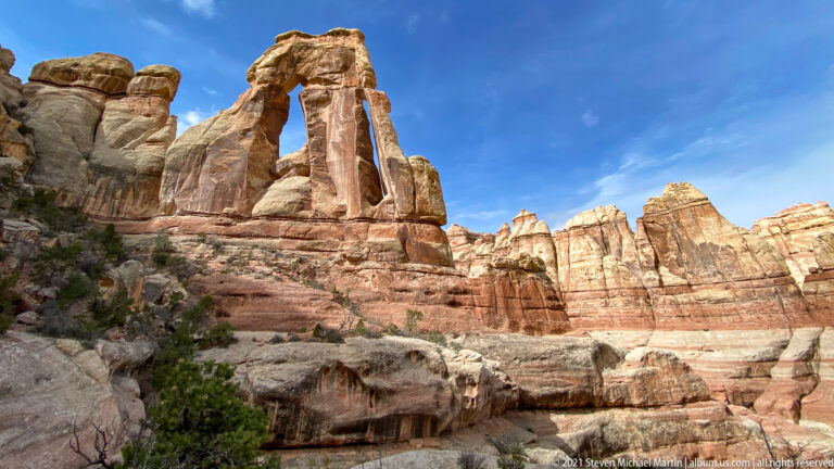 Druid Arch stands against blue sky beside a high sandstone wall and pale sloping ledge.