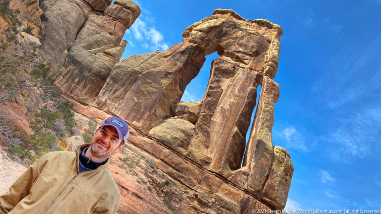 A man in a tan jacket and dark cap poses in the foreground with Druid Arch towering behind him.