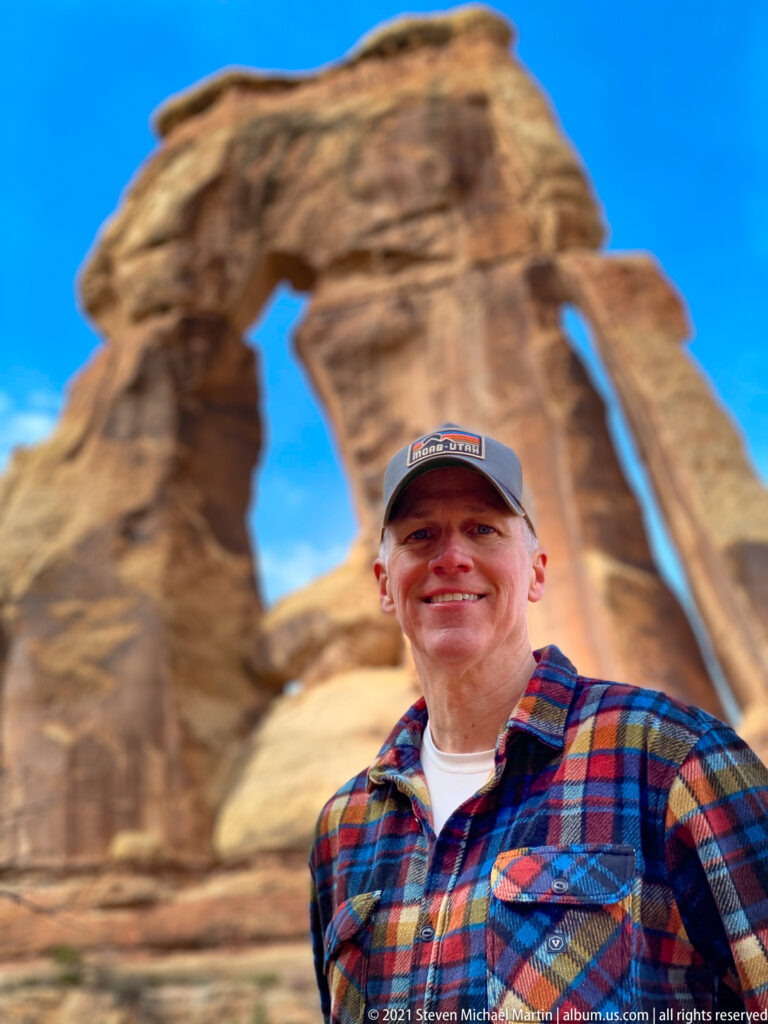 A smiling man in a plaid shirt stands in focus, with Druid Arch blurred behind him.