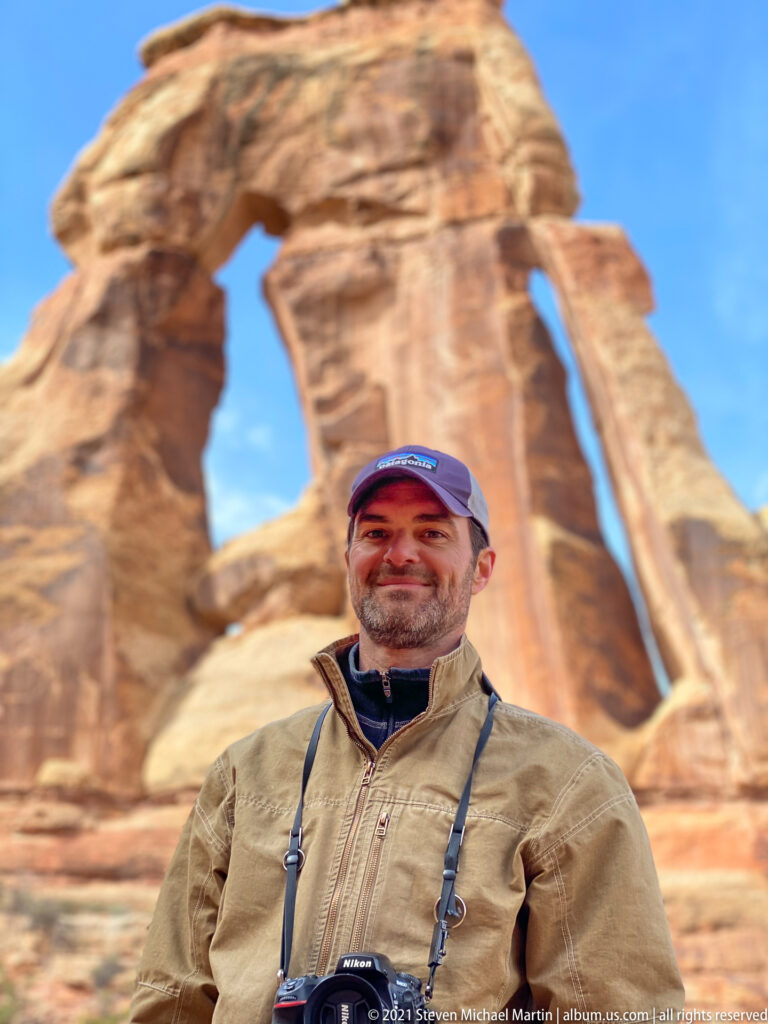 A smiling hiker in a tan jacket and cap stands close to the camera with Druid Arch blurred behind.