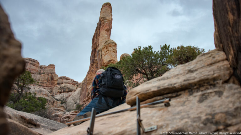 A backpacked hiker climbs stone steps beside a metal handrail, with a narrow sandstone spire behind.