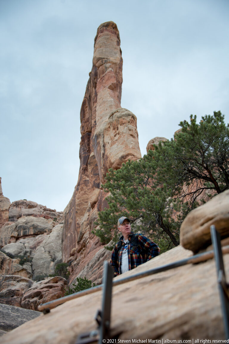 A visitor climbs behind a low stair wall beneath a narrow sandstone tower, with handrails in front.