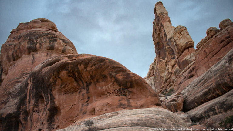 A narrow sandstone pinnacle rises behind a rounded red-rock dome between steep walls.