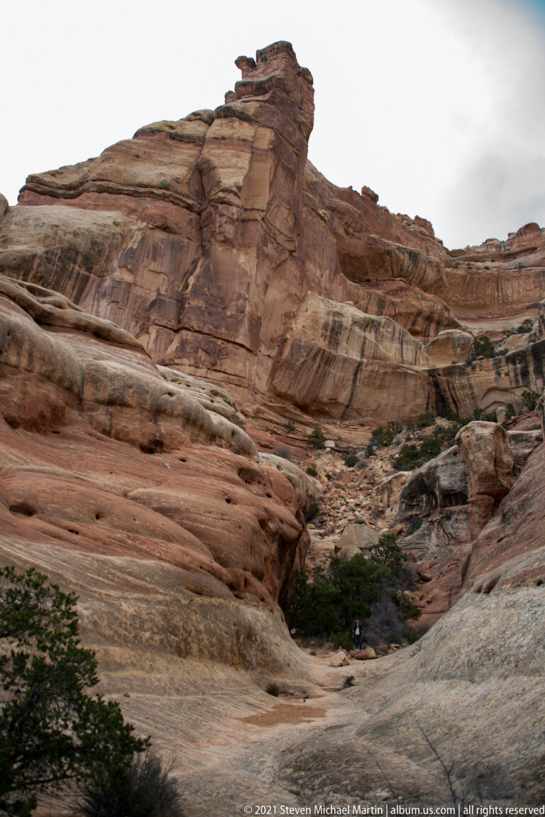 A small hiker stands beneath a massive banded cliff at the end of a narrow rocky passage.