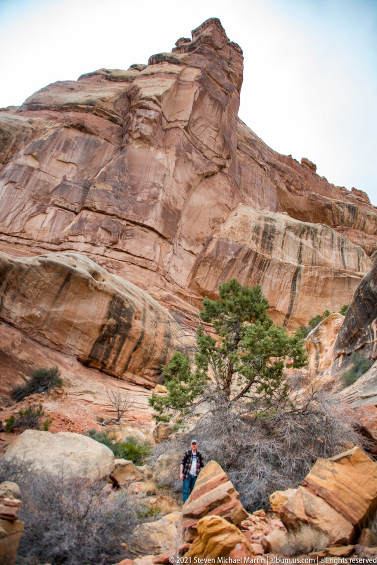 A visitor stands among foreground boulders beneath a tall layered sandstone cliff and a patch of sky.