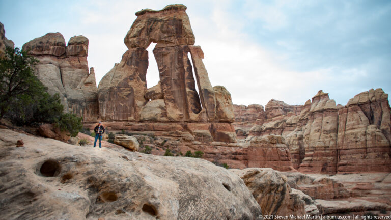 A lone visitor stands on a sloping sandstone ledge below the towering opening of Druid Arch.