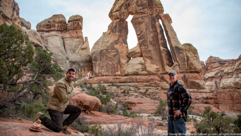 Two visitors pose beneath Druid Arch, with one kneeling beside a boulder and gesturing toward the formation.