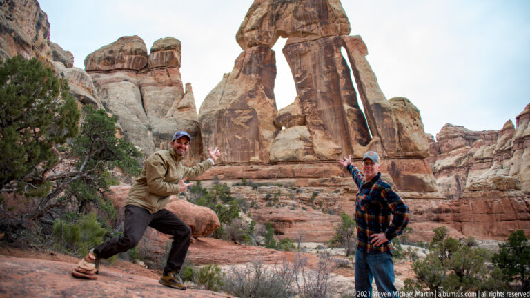 Two visitors raise their hands toward Druid Arch, standing far below its massive sandstone capstone.