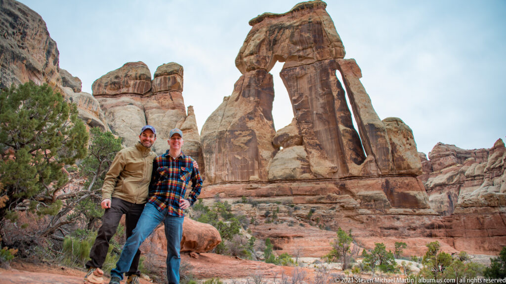 Two visitors stand together on a rocky slope beneath Druid Arch, with juniper and sandstone around them.