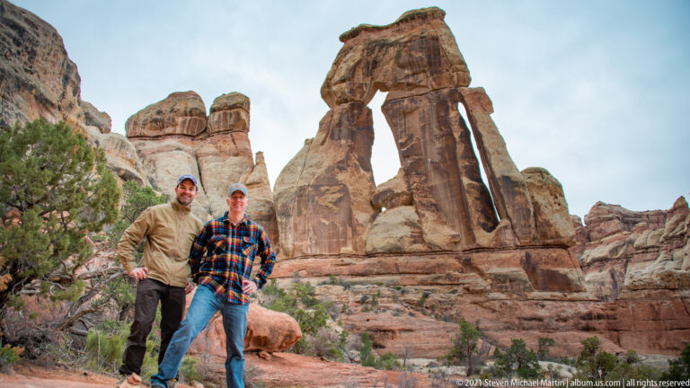 Two visitors pose side by side below Druid Arch, framed by tall sandstone supports and open sky.