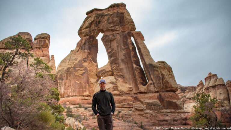 A lone hiker stands below the opening of Druid Arch, surrounded by sandstone and low junipers.