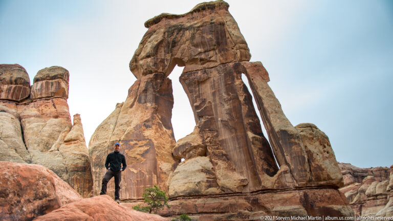 A small hiker stands at the left base of Druid Arch beneath the high sandstone span and bright sky.
