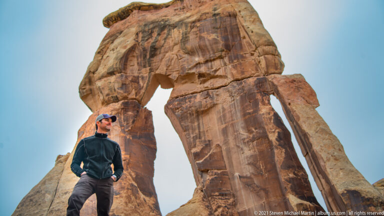 A visitor looks up from below as Druid Arch fills the sky above a tall sandstone support.