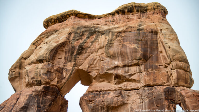 Druid Arch’s broad sandstone capstone spans a large opening, showing vertical streaks and ledges against the sky.