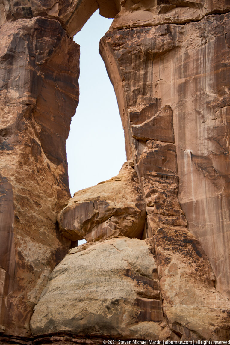 Pale sky shows through Druid Arch, framed by close sandstone walls and layered ledges below.