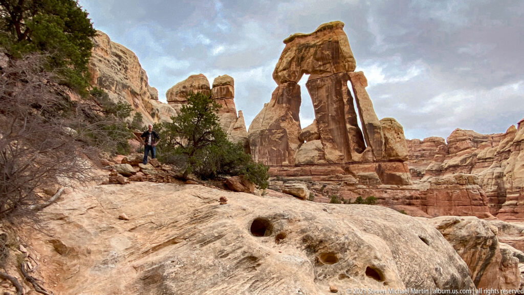 A hiker stands on a pale rock shelf below Druid Arch, with dark depressions in the foreground stone.
