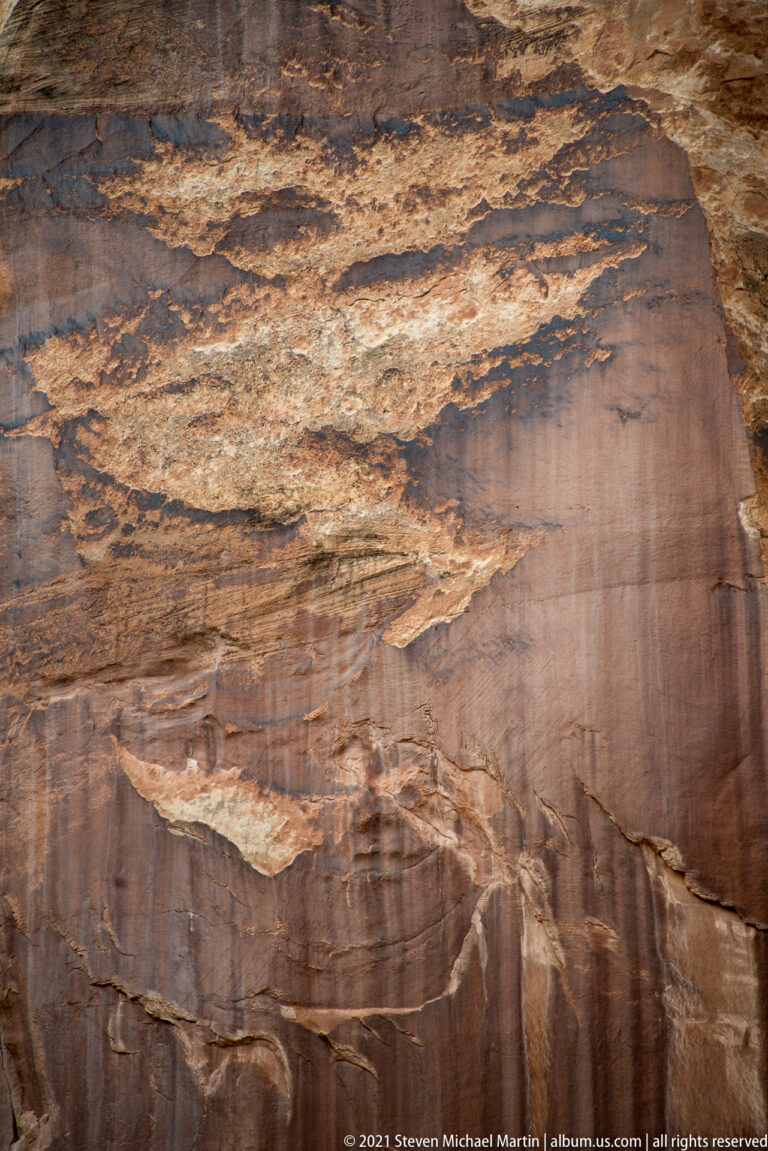 Dark vertical streaks descend across a smooth sandstone wall above thin horizontal layers and ledges.