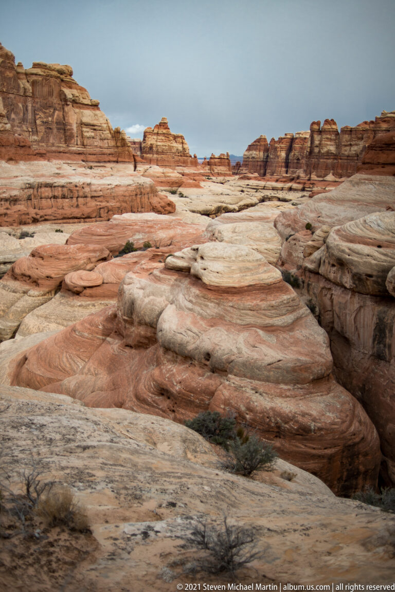 Pale sandstone shelves and shallow channels stretch toward distant layered cliffs, with sparse shrubs on the basin floor.