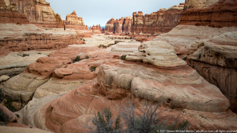 A slickrock corridor leads between pale sandstone shelves toward high red cliffs in the distance.