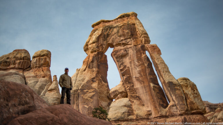 A lone hiker stands on the lower left ledge beside Druid Arch’s towering sandstone support.