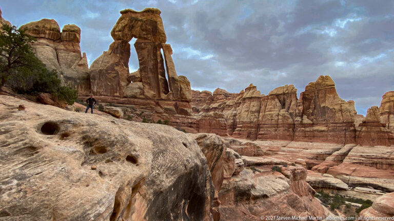 Druid Arch rises above a brushy basin and layered sandstone, with a pitted slab and hiker in front.