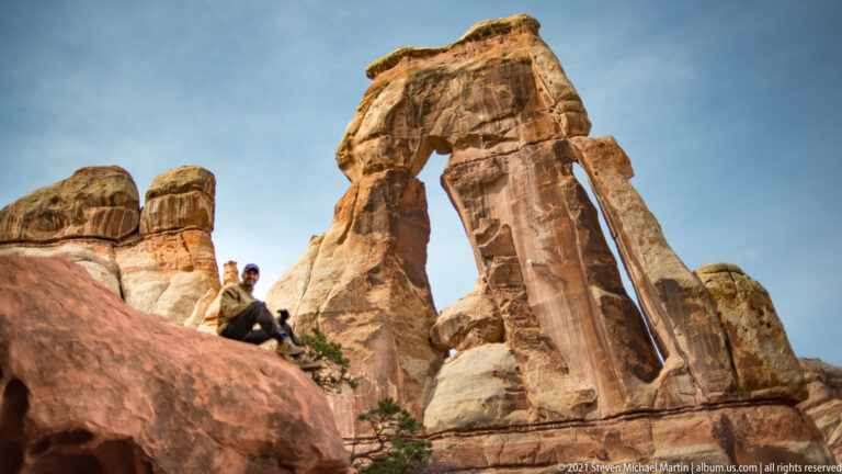 Two visitors sit low on a rocky ledge, partly screened by a boulder beneath Druid Arch.