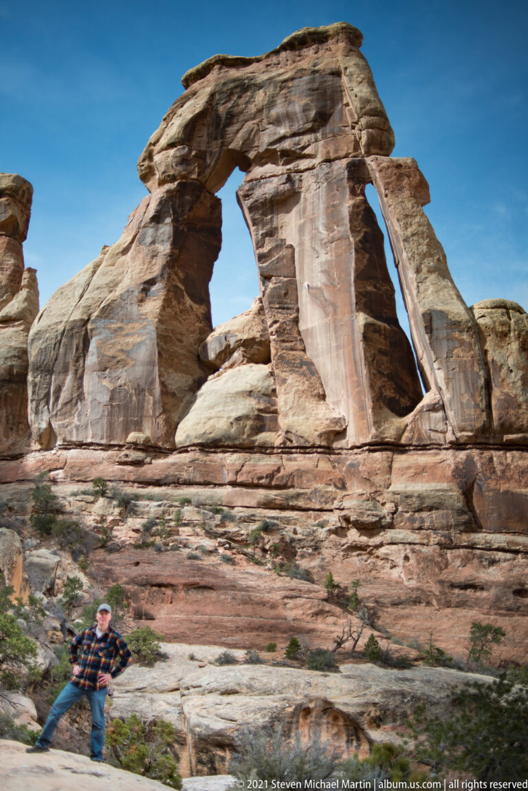 A visitor poses at the far left below Druid Arch, with sandstone benches and blue sky around the span.