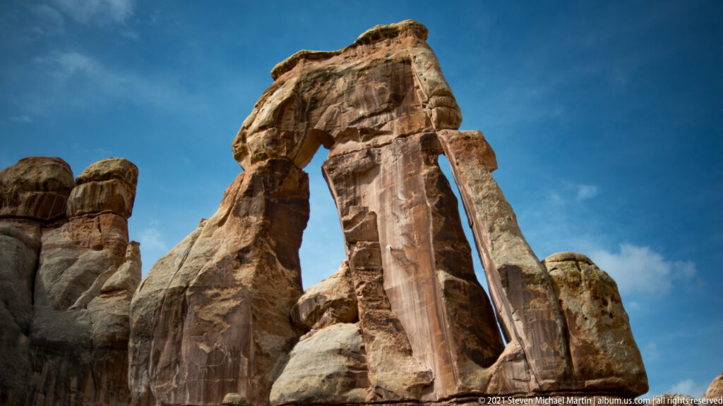 Druid Arch’s uneven capstone bridges tall supports beneath a clear blue sky, with a narrow side opening at right.