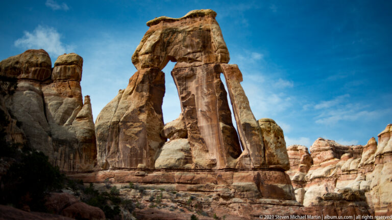 Sunlit Druid Arch rises above a dark foreground shadow, with its openings visible against blue sky.