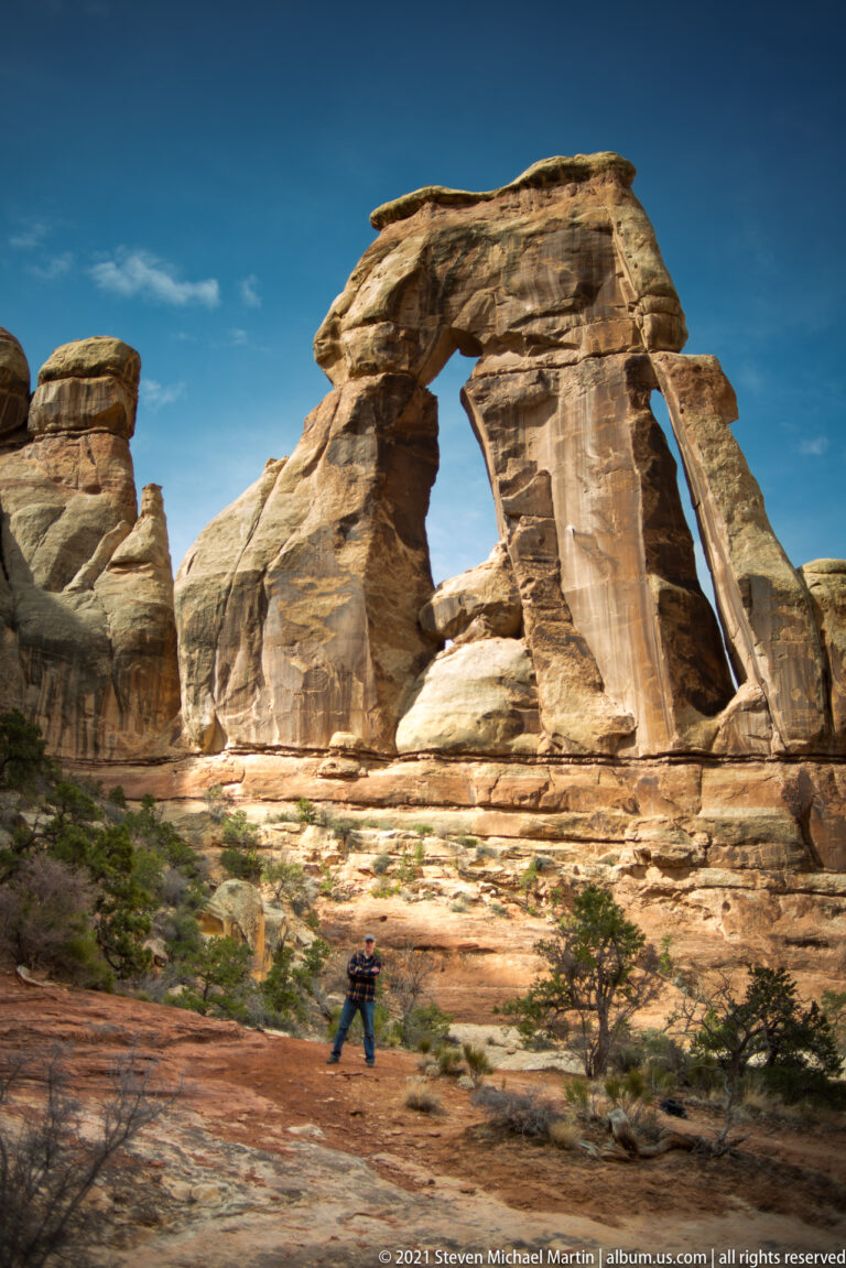 A small visitor stands in the lower center beneath Druid Arch, with canyon walls visible through the opening.