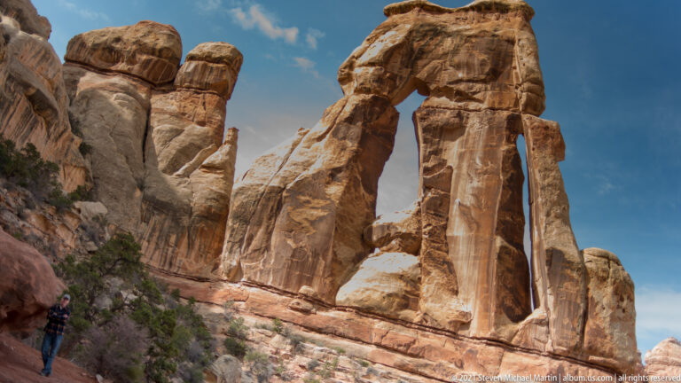 A hiker stands below Druid Arch, a tall sandstone formation with a broad cap and narrow opening beneath a blue sky.