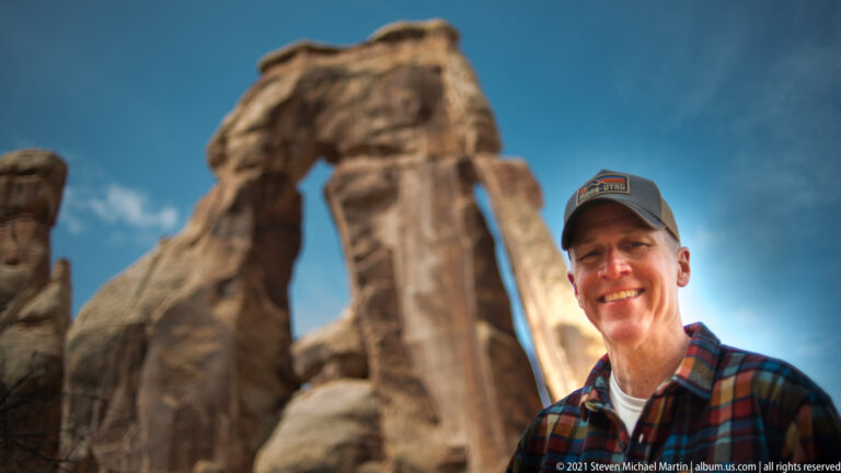 A smiling hiker in a cap and plaid shirt stands before a softly blurred sandstone arch under a blue sky.