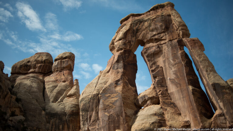 A low-angle view looks up at Druid Arch and neighboring sandstone pinnacles against a blue, lightly clouded sky.