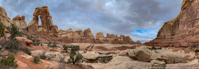 Druid Arch rises at left above a broad canyon floor framed by sandstone towers and a high right-hand wall.
