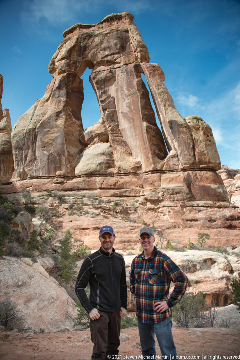 Two hikers pose beneath Druid Arch, framed by the full sandstone span and the rocky ground below.