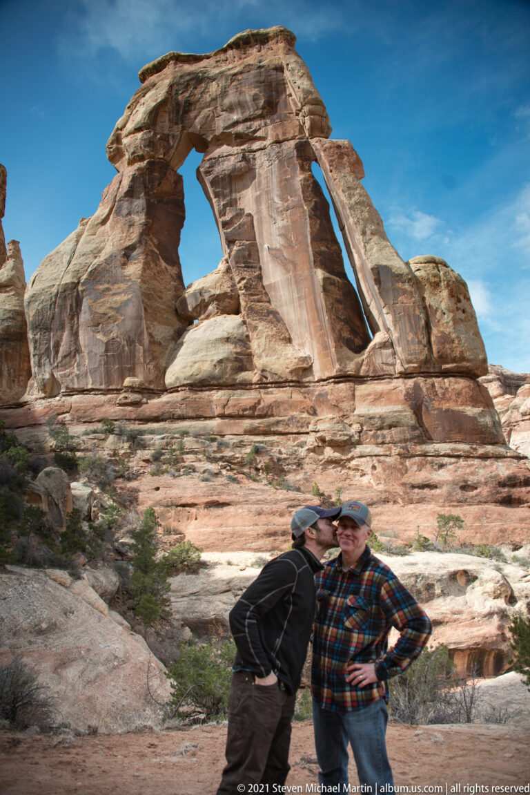 Two hikers stand close together on pale rock, with Druid Arch spanning the background above them.