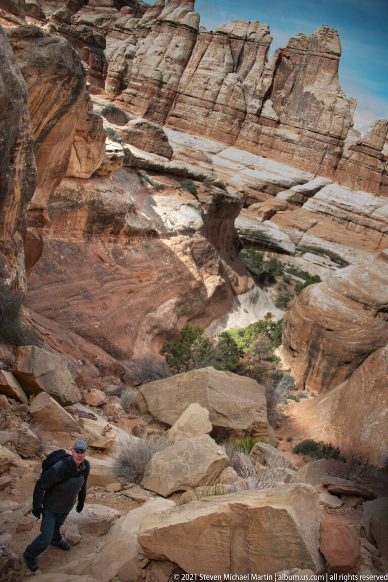 A hiker steps across uneven sandstone beneath high, layered canyon walls, with broken rock slabs along the route.