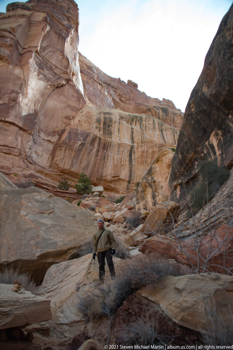 A lone hiker stands among broken sandstone slabs in a drainage enclosed by steep canyon walls.