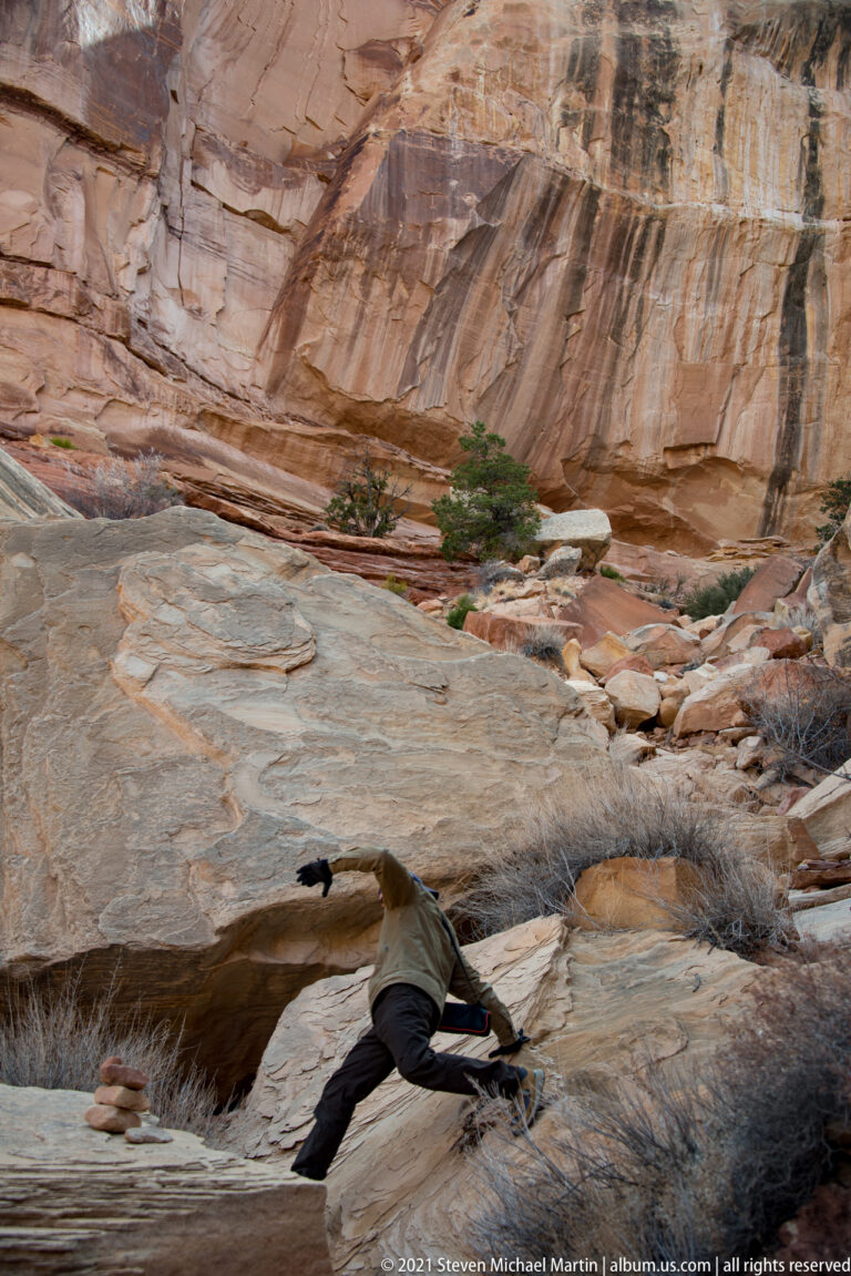 A hiker balances across a gap between sandstone ledges, with a banded canyon wall and shrubs behind.