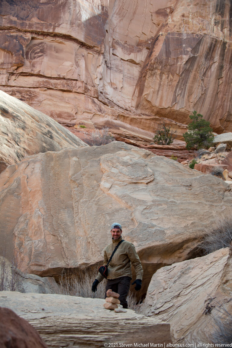 A hiker stands beside a small stone stack among pale slabs beneath a high, banded sandstone wall.